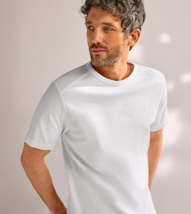 Ein Mann trägt ein weißes T-Shirt und blickt entspannt zur Seite, während er vor einem dezent beleuchteten, neutralen Hintergrund steht.