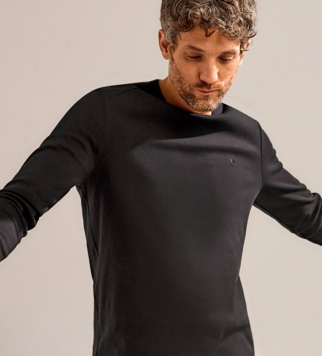 Ein Mann trägt ein schwarzes Langarmshirt und blickt nach unten, während er die Arme seitlich ausstreckt, vor neutralem Hintergrund.
