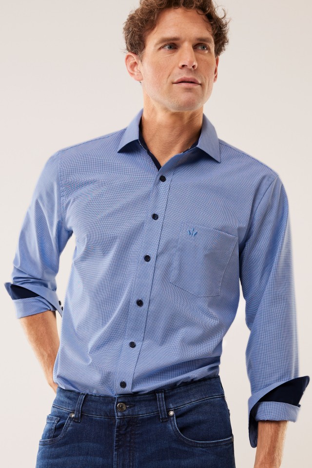 Ein Mann trägt ein hellblaues, fein gemustertes Hemd mit aufgekrempelten Ärmeln und eine dunkelblaue Jeans, während er zur Seite blickt.