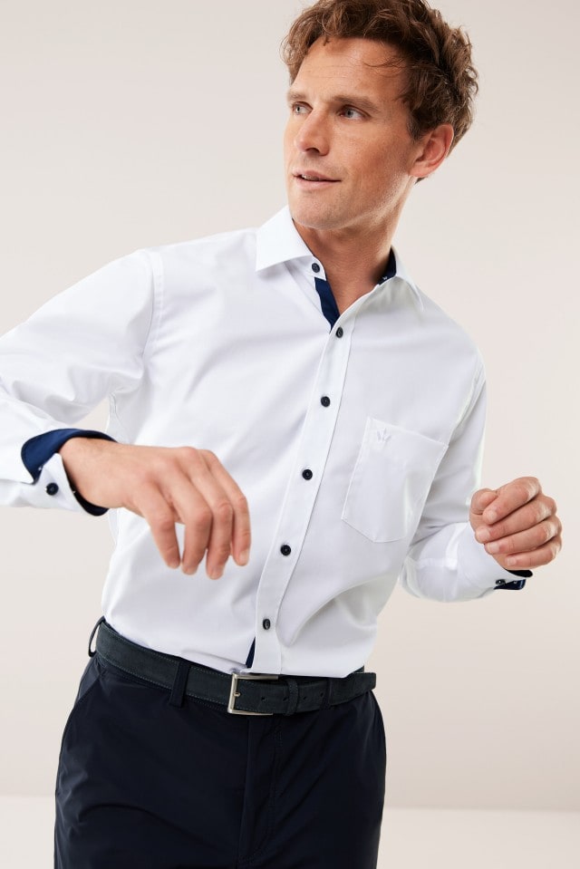 Ein Mann trägt ein weißes Hemd mit dunkelblauen Knöpfen und Innendetails sowie eine dunkle Hose und blickt zur Seite.