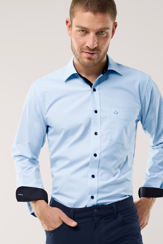 Ein Mann trägt ein hellblau-weiß gestreiftes Hemd mit dunkelblauen Knöpfen und Details sowie eine dunkelblaue Hose und blickt direkt in die Kamera.