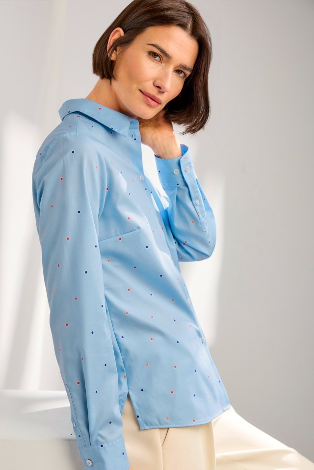 Eine Person trägt eine hellblaue Bluse mit kleinen, farbigen Punkten und kombiniert diese mit einer hellen Hose.