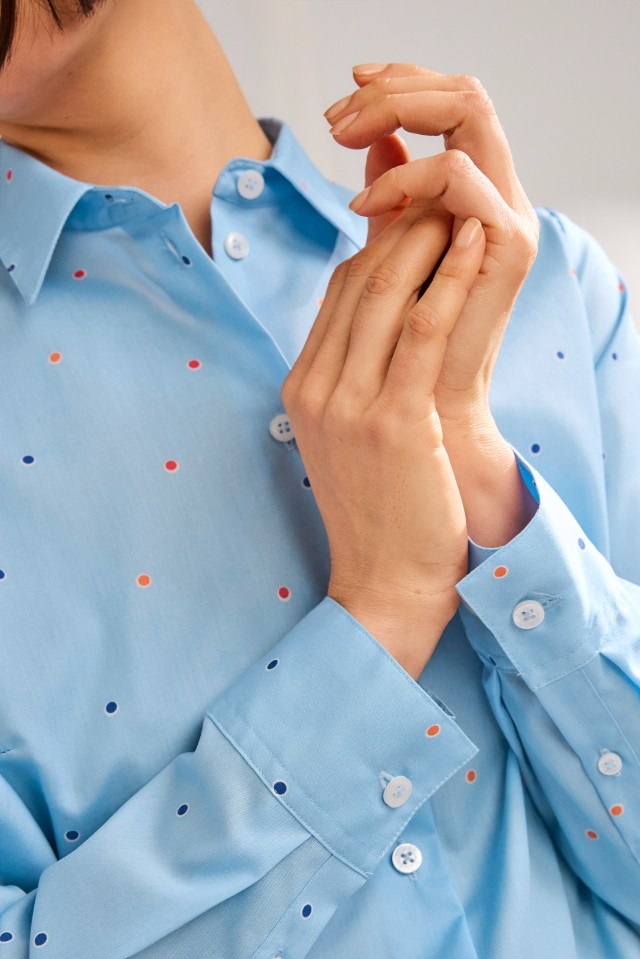 Eine Person trägt eine hellblaue Bluse mit kleinen bunten Punkten und weißen Knöpfen.