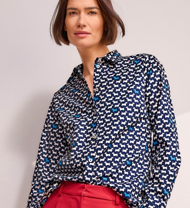 Eine Frau trägt eine langärmlige Bluse mit blau-weißem Hundefiguren-Muster und eine rote Hose, während sie in die Kamera blickt.