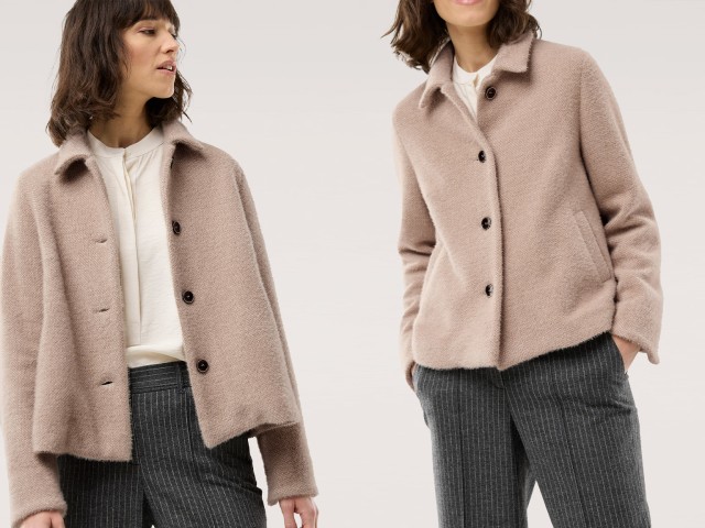 Eine Frau trägt einen beigen Kurzblazer mit Knöpfen über einer weißen Bluse. Dazu kombiniert sie eine dunkelgraue Bundfaltenhose mit feinen Nadelstreifen für einen eleganten Business-Look.