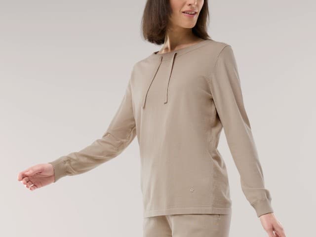 Eine Frau trägt ein beiges Langarmshirt mit Bindeband am Halsausschnitt und passender Hose. Der Look ist bequem und wirkt harmonisch Ton in Ton.