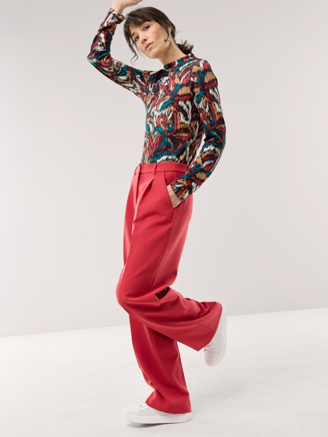 Eine Frau trägt eine weite, leuchtend rote Hose mit Bundfalten und weißen Sneakern. Dazu kombiniert sie ein bunt gemustertes, langärmliges Oberteil für einen modernen und ausdrucksstarken Look.