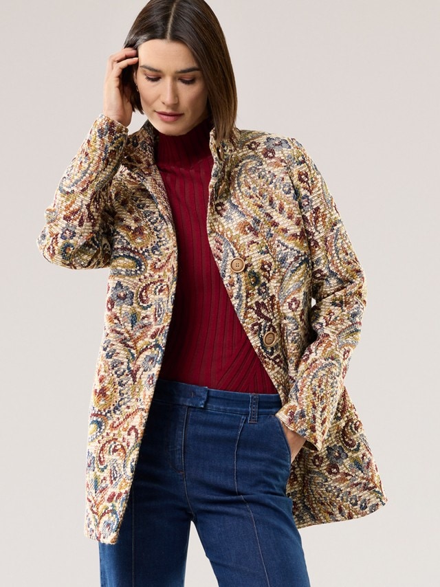 Eine Frau trägt eine bunt gemusterte Jacke im Paisley-Stil über einem roten Strickpullover. Dazu kombiniert sind schlichte blaue Jeans für einen eleganten, aber alltagstauglichen Look.
