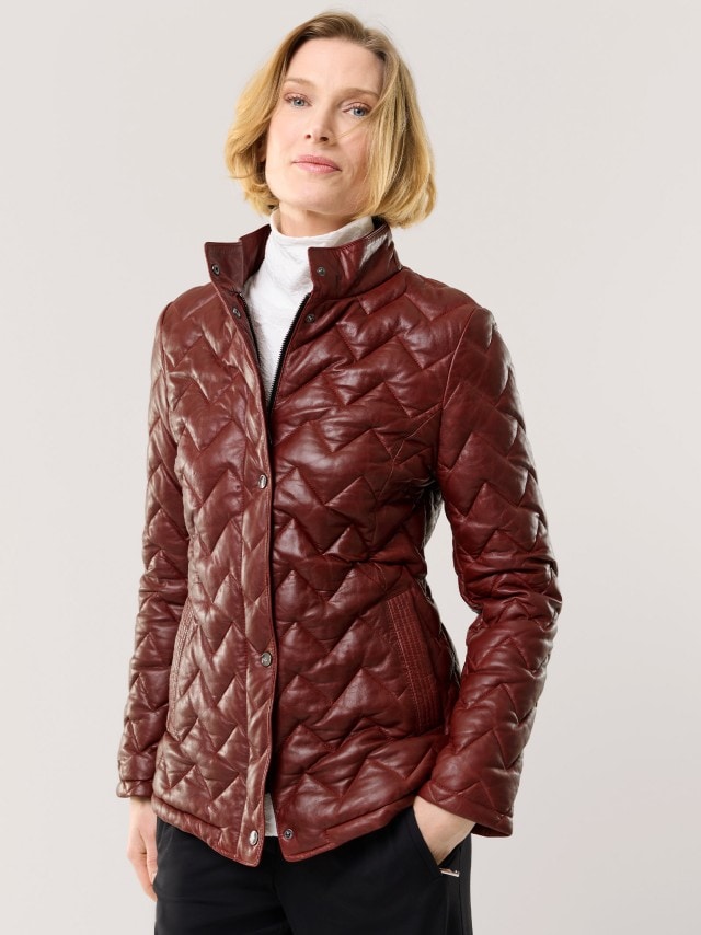 Eine Frau trägt eine gesteppte, weinrote Jacke aus Lederoptik über einem weißen Rollkragenpullover. Der Look wirkt elegant und zugleich modern.