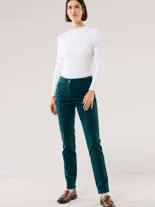 Eine Frau trägt einen weißen Ripppullover mit langen Ärmeln. Dazu kombiniert sie eine smaragdgrüne Hose und braune Loafers für einen frischen, eleganten Look.