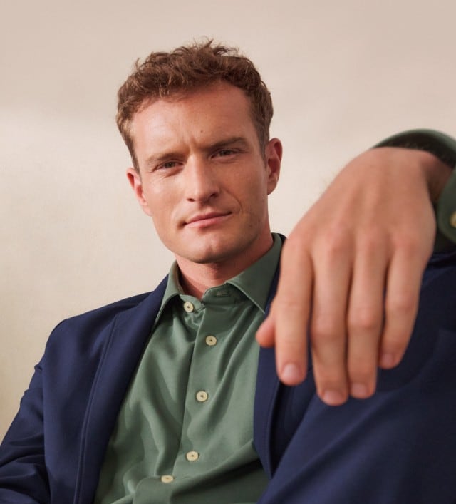 Zwei Männer in modischer Kleidung sitzen entspannt nebeneinander. Einer trägt ein graues, strukturiertes Overshirt mit Rollkragenpullover, der andere einen dunkelblauen Anzug mit grünem Hemd.