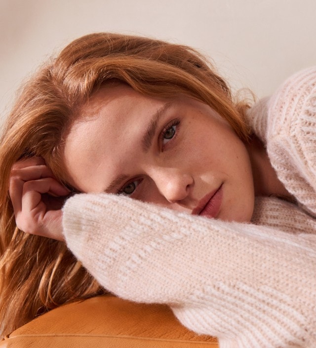 Eine Frau liegt entspannt und trägt einen weichen, cremefarbenen Strickpullover mit dezentem Muster. Der kuschelige Look wirkt gemütlich und lässig.