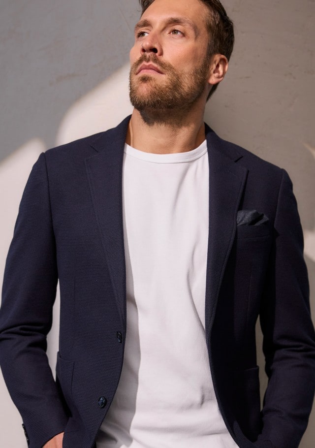 Ein Mann trägt ein dunkelblaues Sakko, darunter ein weißes Shirt. 