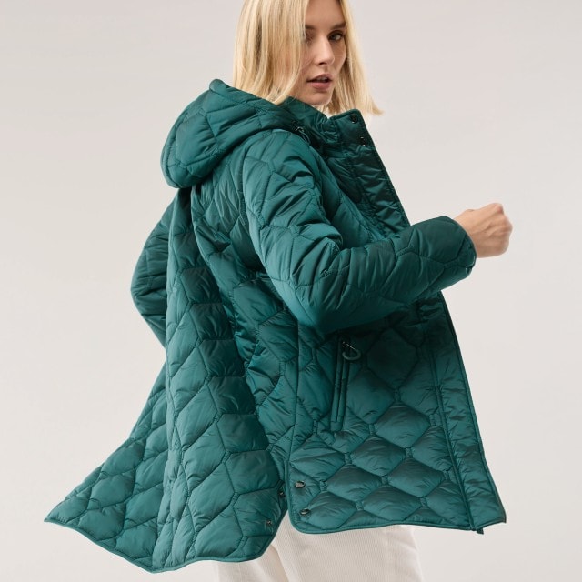 Eine Frau trägt eine gesteppte, smaragdgrüne Winterjacke mit Kapuze. Der Look wirkt modern und sportlich-elegant.