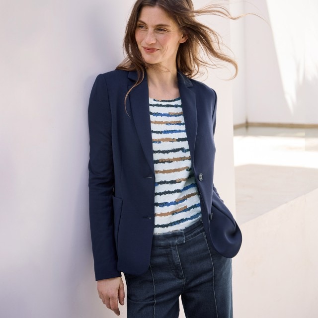 Eine Frau trägt einen dunkelblauen Blazer über einem gestreiften Shirt in Blau-, Beige- und Weißtönen. Der Look ist casual-elegant und wirkt modern.