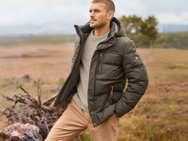 Mann in einem hellen Strickpullover, hellbrauner Hose und olivgrüner Steppjacke steht in einem Feld und schaut zur Seite.