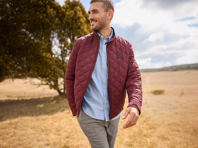 Ein Mann in einem hellblauen Hemd, rostroter Steppjacke und gemusterter Hose läuft über ein Feld und schaut lächelnd zur Seite.