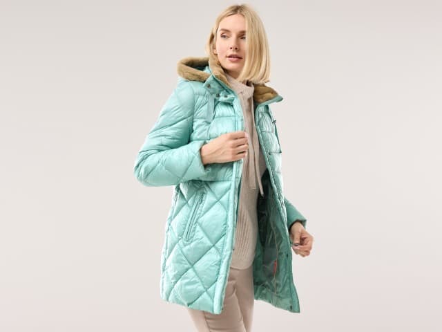 Eine Frau trägt die eine Steppjacke mit Kuschelkragen in Gletschergrün, darunter einen Rollkragen-Strickpullover in Creme und dazu eine Hose in Beige.