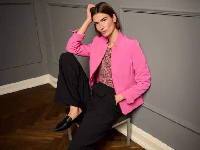 Eine Frau sitzt lässig auf einem Stuhl und trägt den Kofferblazer Airline in Soft Pink, darunter ein Viskoseshirt in Bouclelook und dazu eine schwarze Hose. 