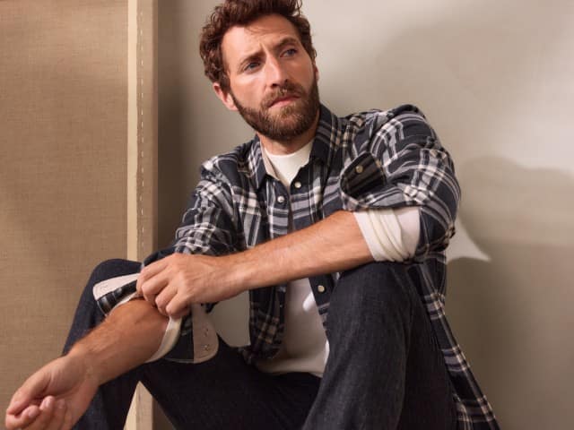 Ein Mann in einem schwarz-weiß karierten Flanellhemd über einem weißen Rollkragenpullover und Jeans sitzt vor einer Wand und schaut zur Seite.