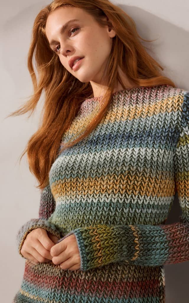 Ein Frau schaut zur Seite und trägt einen Grobstrick-Pullover mit Farbverlauf in Oliv/Siena.