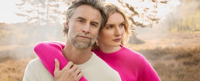 Ein Mann und eine Frau stehen eng nebeneinander in der Natur. Sie tragen Pullover, der Mann in Creme und die Frau in Pink.