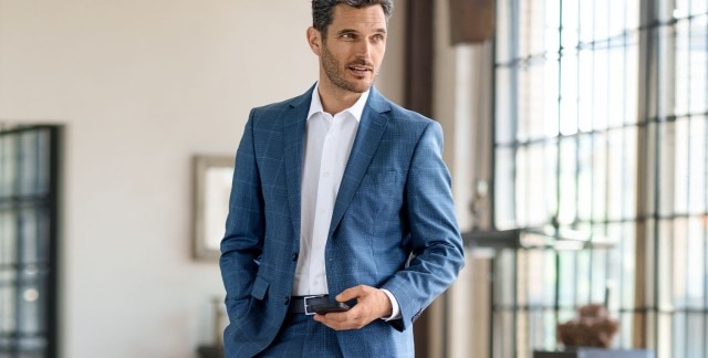 Mann steht in einem Loft und trägt einen blau karierten Anzug und hält ein Handy in der Hand.