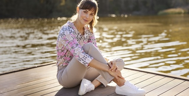 Frau mit gemusterter Bluse und beigefarbener Hose sitzt auf einem Steg am Wasser