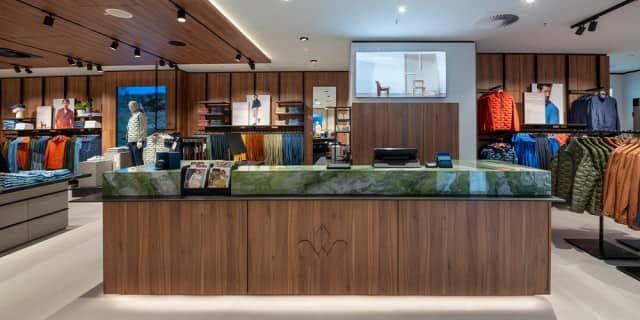 Das Bild zeigt den Innenraum eines Walbusch-Geschäfts mit einer modernen Holz-Verkaufstheke im Vordergrund und ordentlich präsentierter Kleidung auf Regalen und Kleiderständern im Hintergrund.