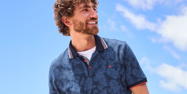 Ein Mann trägt ein dunkelblaues Poloshirt mit feinem floralen Muster und roten Akzenten am Kragen und den Knöpfen. Der Look ist sommerlich, modern und wirkt entspannt – perfekt für sonnige Tage im Freien.