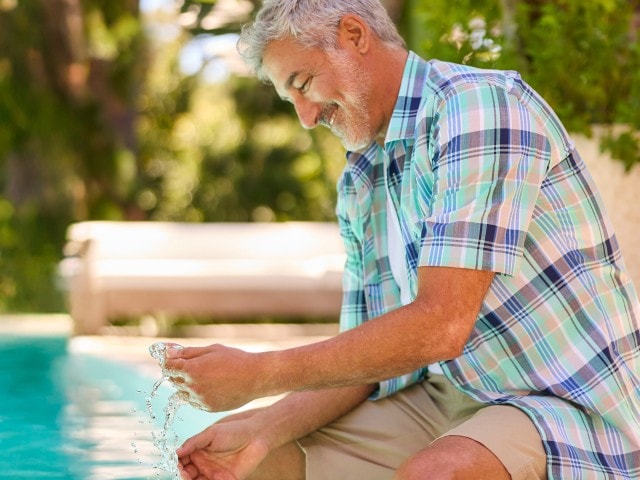 Ein Mann sitzt am Pool, spielt mit Wasser und trägt ein kurzärmliges, kariertes Hemd in verschiedenen Blau-, Grün- und Weißtönen sowie eine beige Shorts.