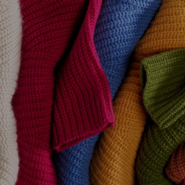 Gestrickte Stoffrollen in fünf Farben: Weiß, Rot, Blau, Gelb und Grün, eng nebeneinander angeordnet.