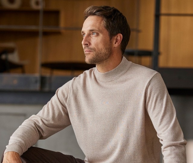 Herr sitzt und trägt einen beigen Merino Rolkragen Pullover.