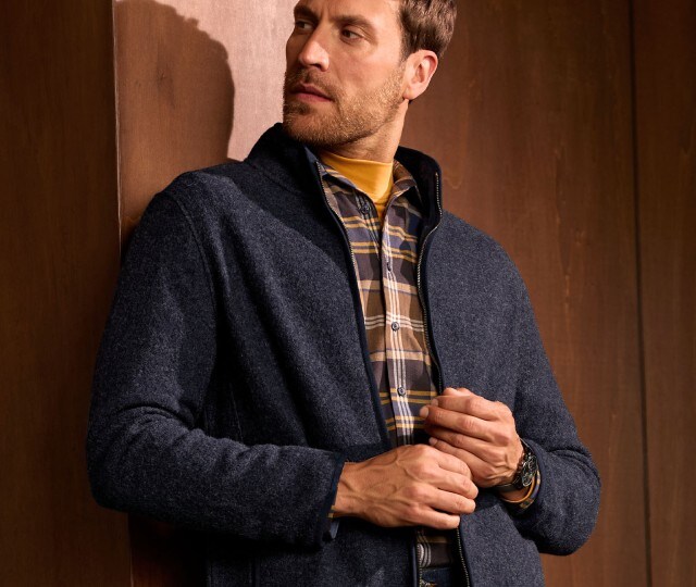 Herr lehnt an einer Wand und trägt eine blaue Strickjacke mit Karo Hemd.