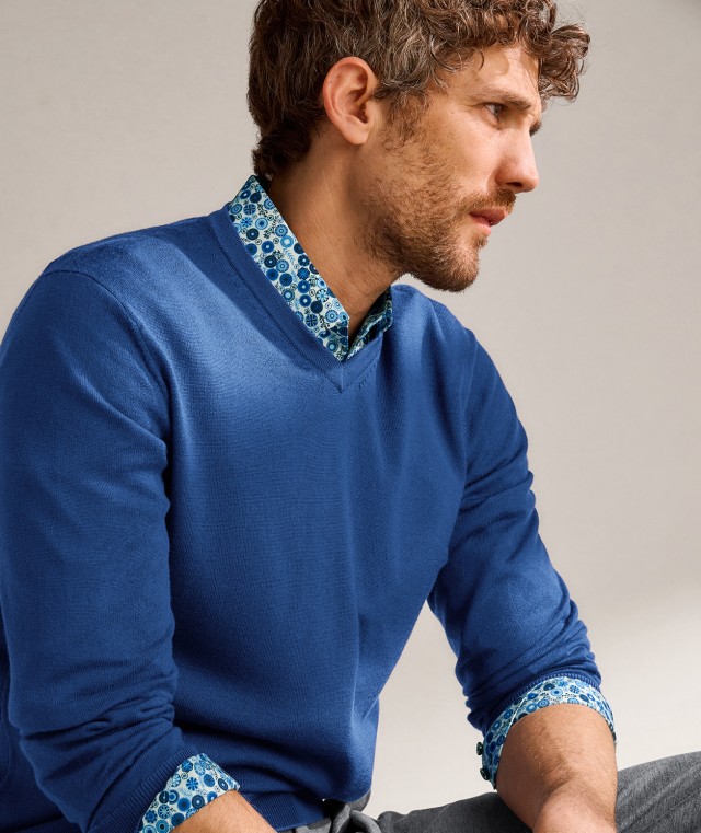 Ein Mann trägt einen dunkelblauen Pullover mit Rundhalsausschnitt, darunter ein gemustertes Hemd.