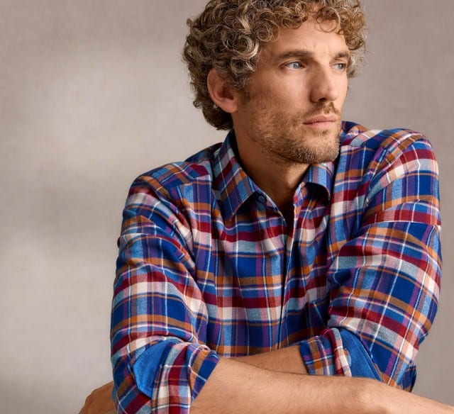 Ein Mann mit lockigem Haar, trägt ein Flanellhemd und schaut zur Seite.
