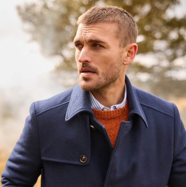 Das Bild zeigt einen Mann, der im Freien steht und einen dunkelblauen Mantel, einen orangefarbenen Pullover und ein hellblau-weiß gestreiftes Hemd trägt.