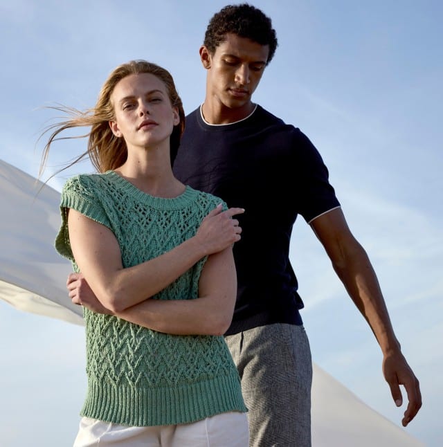 Zwei Personen stehen im Freien vor einem hellen Himmel; die Frau trägt ein mintgrünes, gestricktes Oberteil und weiße Hose, der Mann ein dunkelblaues T-Shirt mit heller Kante und graue Hose. Ihr Stil wirkt modern, luftig und sommerlich, passend zu einer natürlichen, eleganten Atmosphäre.