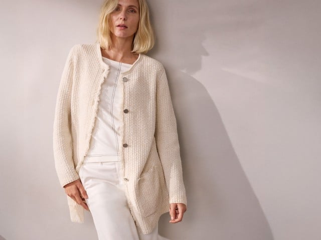 Eine Frau trägt eine cremefarbene Strickjacke über einem weißen Outfit und lehnt an einer Wand. Eine Frau trägt eine cremefarbene Strickjacke über einem weißen Outfit und lehnt an einer Wand.