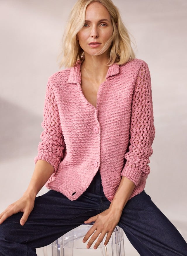 Eine Frau trägt eine rosafarbene Strickjacke mit durchbrochenen Ärmeln und sitzt auf einem transparenten Hocker. Eine Frau trägt eine rosafarbene Strickjacke mit durchbrochenen Ärmeln und sitzt auf einem transparenten Hocker.