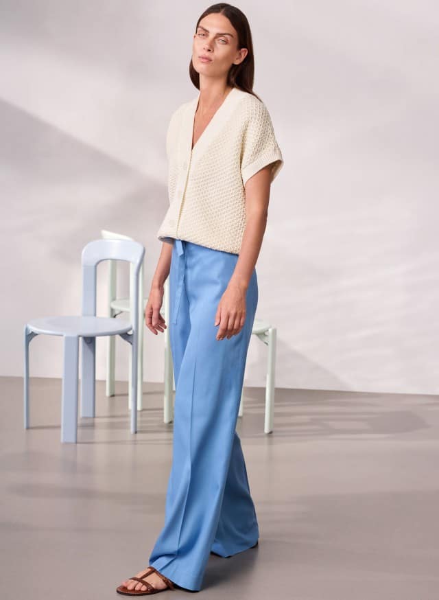 Eine Frau trägt eine cremefarbene kurze Strickjacke und eine weite hellblaue Hose und geht durch einen minimalistisch eingerichteten Raum. Eine Frau trägt eine cremefarbene kurze Strickjacke und eine weite hellblaue Hose und geht durch einen minimalistisch eingerichteten Raum.