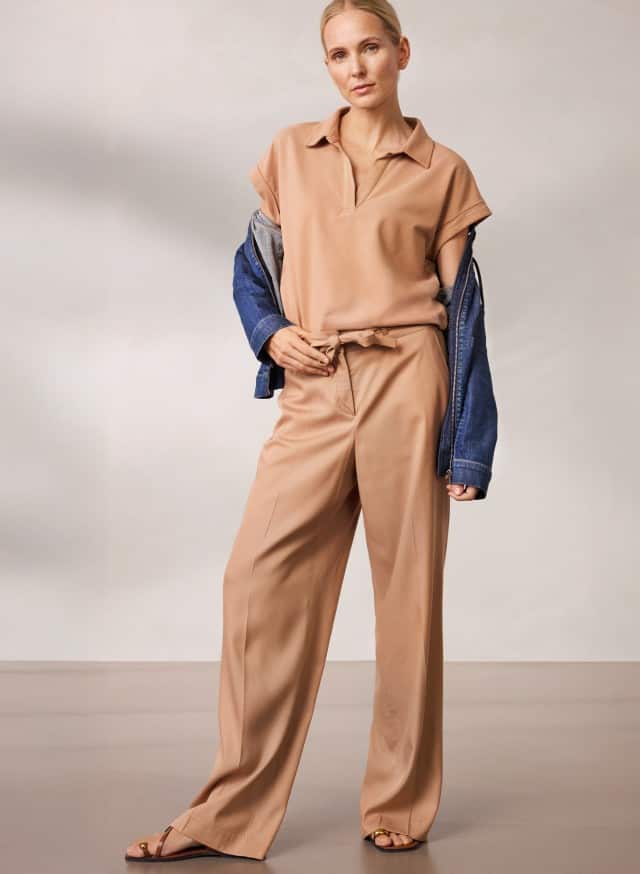 Eine Person steht vor einer neutralen Wand und trägt ein beigefarbenes Outfit mit weiter Hose und Kurzarmtop, kombiniert mit einer halb übergezogenen Jeansjacke und offenen Sandalen. Eine Person steht vor einer neutralen Wand und trägt ein beigefarbenes Outfit mit weiter Hose und Kurzarmtop, kombiniert mit einer halb übergezogenen Jeansjacke und offenen Sandalen.