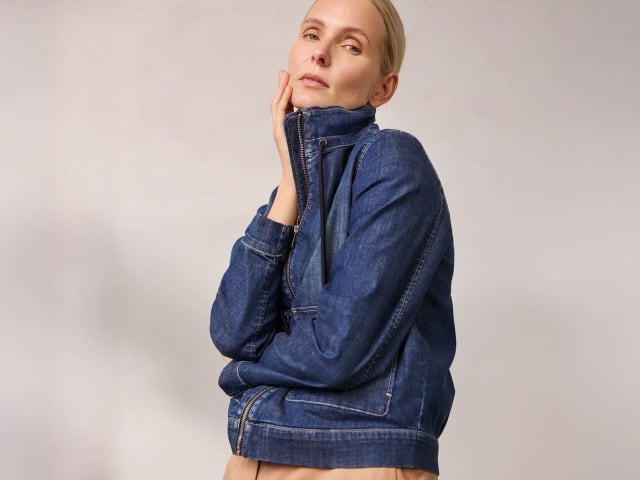 Eine Person trägt eine hochgeschlossene dunkelblaue Jeansjacke und berührt mit einer Hand ihr Gesicht. Eine Person trägt eine hochgeschlossene dunkelblaue Jeansjacke und berührt mit einer Hand ihr Gesicht.