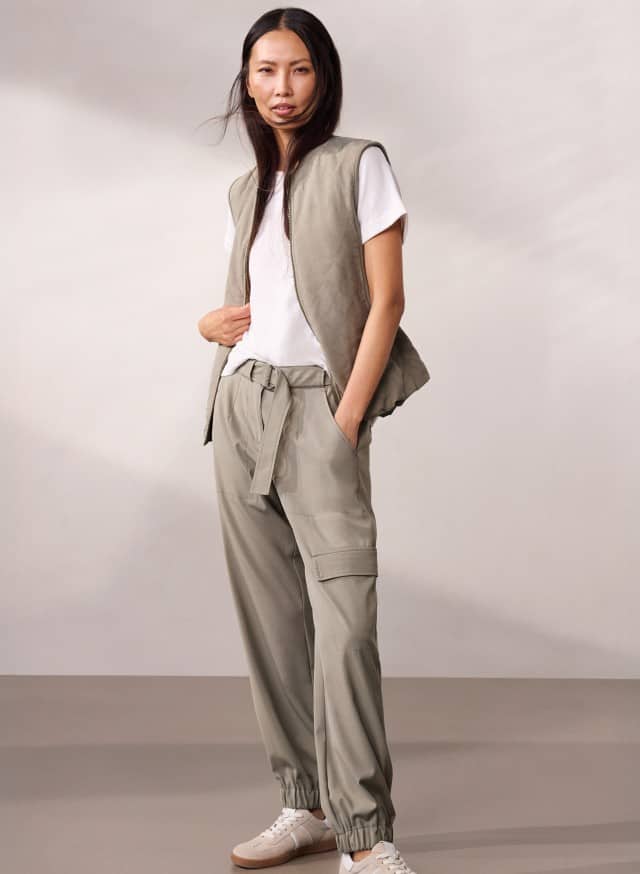 Eine Person steht vor einer neutralen Wand und trägt eine weiße T‑Shirt‑Weste‑Kombination mit locker sitzender, beigefarbener Cargohose und Sneakers. Eine Person steht vor einer neutralen Wand und trägt eine weiße T‑Shirt‑Weste‑Kombination mit locker sitzender, beigefarbener Cargohose und Sneakers.