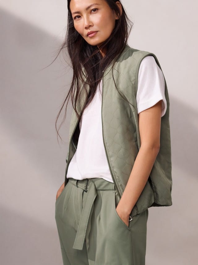 Eine Frau trägt eine leichte, gesteppte Weste zu weißem T‑Shirt und locker gebundenen Stoffhosen, was entspannt, modern und angenehm lässig wirkt. Eine Frau trägt eine leichte, gesteppte Weste zu weißem T‑Shirt und locker gebundenen Stoffhosen, was entspannt, modern und angenehm lässig wirkt.