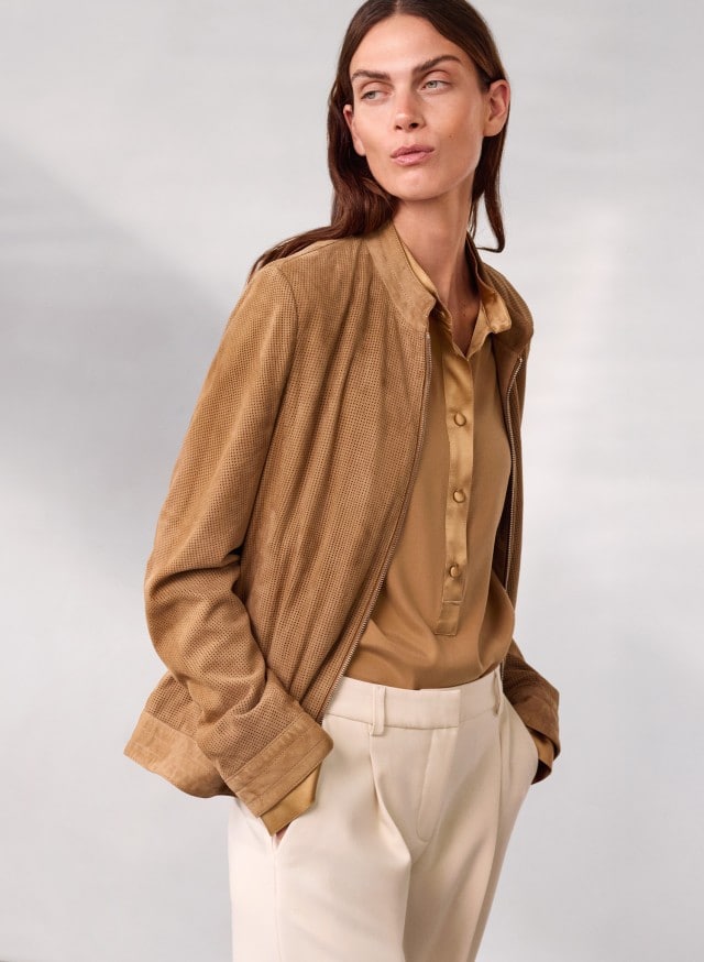 Eine Person trägt eine hellbraune Bluse, eine passende braune Jacke und eine helle Stoffhose und blickt zur Seite. Eine Person trägt eine hellbraune Bluse, eine passende braune Jacke und eine helle Stoffhose und blickt zur Seite.