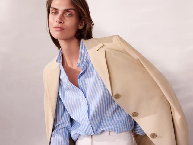 Eine Person trägt eine hellbeige Jacke über einer hellblau‑weiß gestreiften Bluse und einer weißen Hose und lehnt sich leicht nach vorn. Eine Person trägt eine hellbeige Jacke über einer hellblau‑weiß gestreiften Bluse und einer weißen Hose und lehnt sich leicht nach vorn.