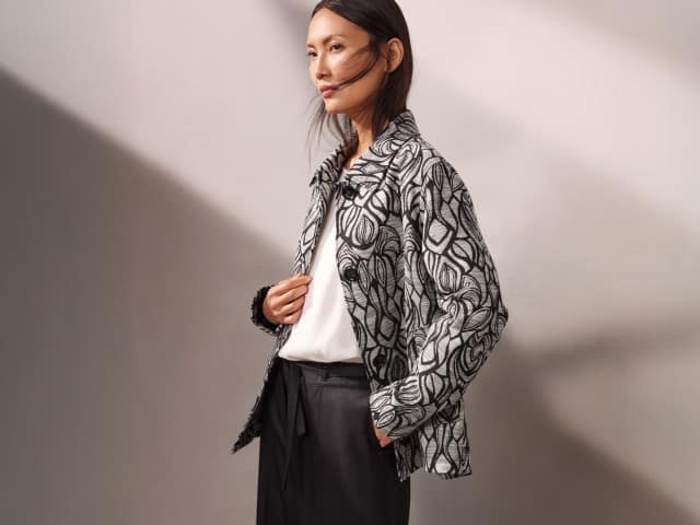 Eine Person trägt eine weiß‑schwarze gemusterte Jacke über einem weißen Oberteil und einer dunklen Hose und blickt zur Seite. Eine Person trägt eine weiß‑schwarze gemusterte Jacke über einem weißen Oberteil und einer dunklen Hose und blickt zur Seite.