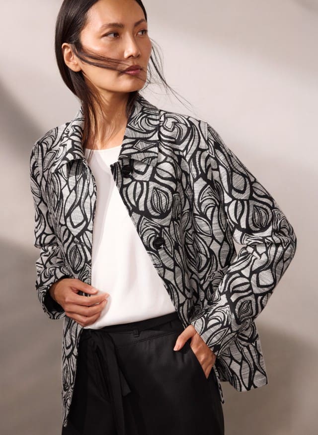 Eine Person trägt eine schwarz‑weiß gemusterte Jacke über einem weißen Oberteil und einer schwarzen Hose und blickt leicht zur Seite. Eine Person trägt eine schwarz‑weiß gemusterte Jacke über einem weißen Oberteil und einer schwarzen Hose und blickt leicht zur Seite.