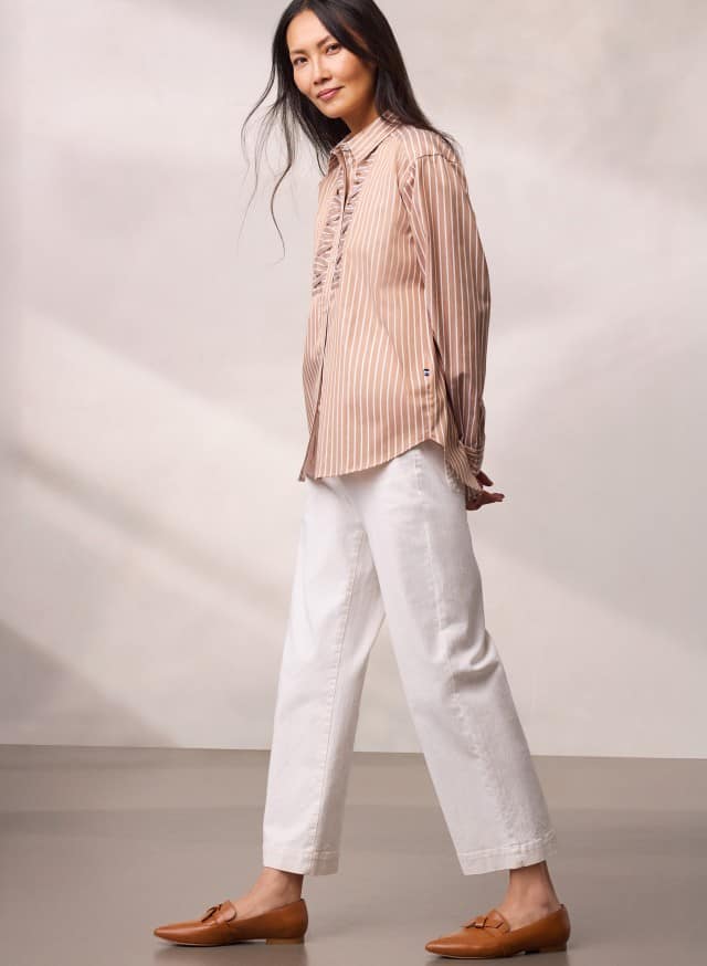 Eine Person trägt eine beigefarbene, gestreifte Bluse zu weißen, weiten Hosen und braunen Loafern und geht leicht zur Seite. Eine Person trägt eine beigefarbene, gestreifte Bluse zu weißen, weiten Hosen und braunen Loafern und geht leicht zur Seite.